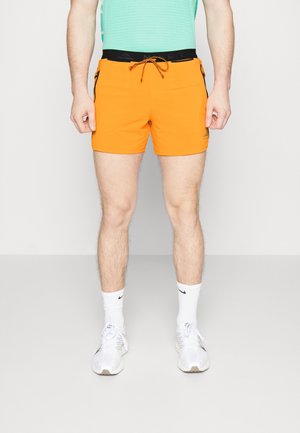 Mann trägt leuchtend orangefarbene Sportshorts mit schwarzem Bund und Kordelzug, weiße Socken und weiße Laufschuhe, stehend vor einem schlichten Hintergrund.