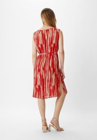 Rot-beige gemustertes Kleid mit ärmellosem Design, ausgestelltem Rock und subtiler Seitenöffnung, aus leichtem Stoff. Sandalen mit quadratischen Absätzen.