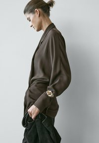 Frau mit hochgestecktem Haar, trägt eine lockere braune Bluse mit metallischem Manschettendetail, hält ein schwarzes Kleidungsstück vor einem hellen, schlichten Hintergrund.