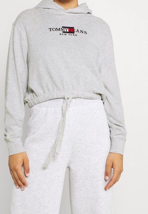 Lichtgrijze cropped hoodie en bijpassende joggingbroek, met "Tommy Jeans New York" logo op de borst, taille met trekkoord, handen rustend langs het lichaam.