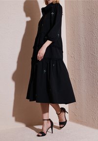 Robe noire à volants avec des manches bouffantes et des broderies florales. Portée avec des talons hautes noirs à lanières. Fond texturé beige.