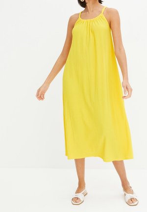 Femme portant une robe midi jaune vif sans manches avec de fines bretelles et des sandales blanches plates, debout devant un fond uni.