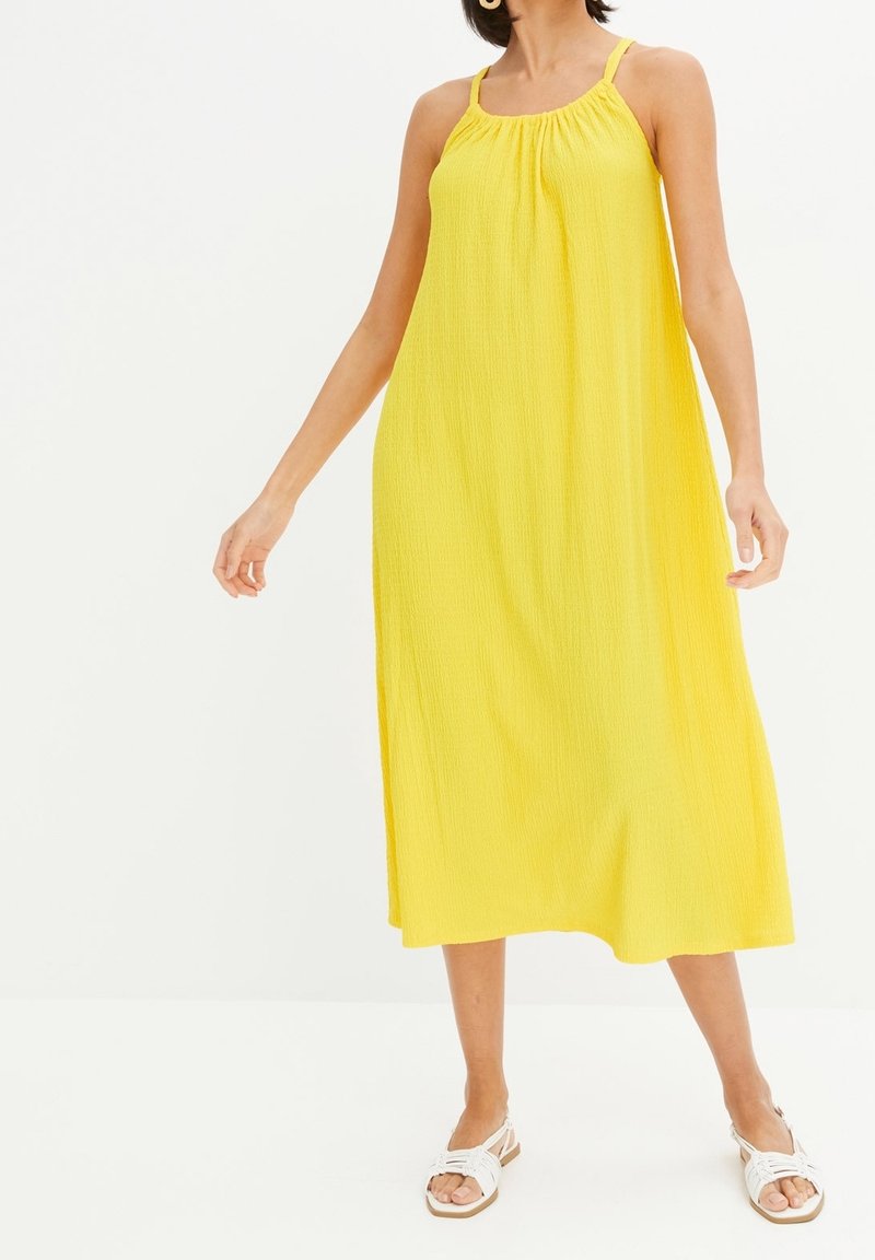 Femme portant une robe midi jaune vif sans manches avec de fines bretelles et des sandales blanches plates, debout devant un fond uni.