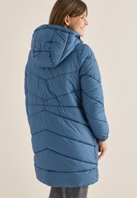 Giacca a vento blu lunga con cappuccio, caratterizzata da motivi a chevron trapuntati, polsini elasticizzati e una vestibilità leggermente rilassata. Materiale leggero.