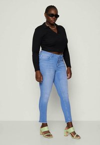 Čierny dlhý rukáv cropped top, svetlomodré úzke džínsy a zelené sandále na blokovom opätku. Model má na sebe tmavé slnečné okuliare a strieborný náhrdelník.