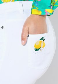 Jeans in denim bianco con una tasca decorata con design di limone giallo ricamato e piccoli strass, che mostrano una texture liscia e una forma aderente.