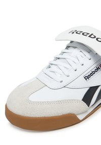 Baltie sneki ar zamša akcentiem un melnu svītru, ar garu mēli un Reebok zīmolu. Brūna gumijas zole. Saistīta dizaina.