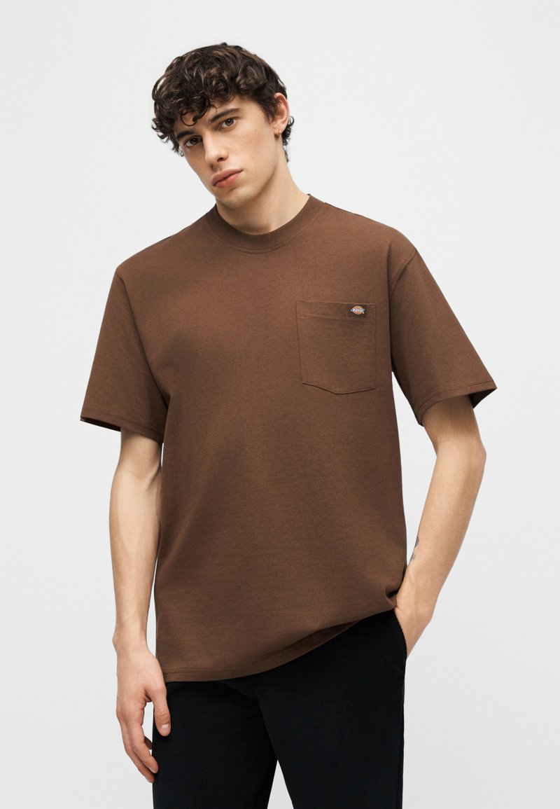 Giovane uomo con capelli ricci che indossa una maglietta marrone a maniche corte con taschino sul petto e pantaloni neri su uno sfondo semplice.