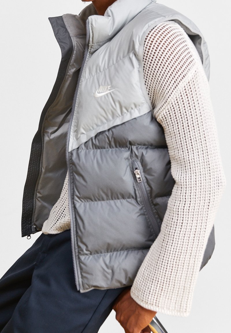 Grå, vadderad Nike-väst med hög krage, dragkedja och sidofickor; har en färgblockerad design och lättviktigt material.