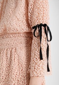 Blusa in pizzo di un rosa tenue con motivo floreale intricato, caratterizzata da maniche lunghe strette con nastri neri e una vita elastica.