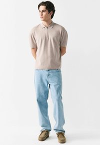 Beige kortærmet polo med lynlås foran, parret med lyseblå jeans og brune sko, stående foran en hvid baggrund.