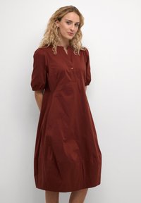 Šaty v hnedej farbe s dizajnom po kolená, krátkymi naberanými rukávmi, bezgolierovým výstrihom a skrytou gombíkovou légou. Hladká, ľahká tkanina.