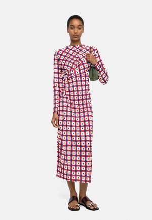 Kobieta stoi, nosząc długą sukienkę midi z geometrycznym wzorem w czerwonym, białym i niebieskim kolorze, trzyma małą zieloną torebkę i nosi czarne sandały.