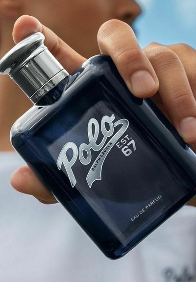 Hand som håller en blå Polo Ralph Lauren parfymflaska med silverlock och text, mot en suddig ljus bakgrund.