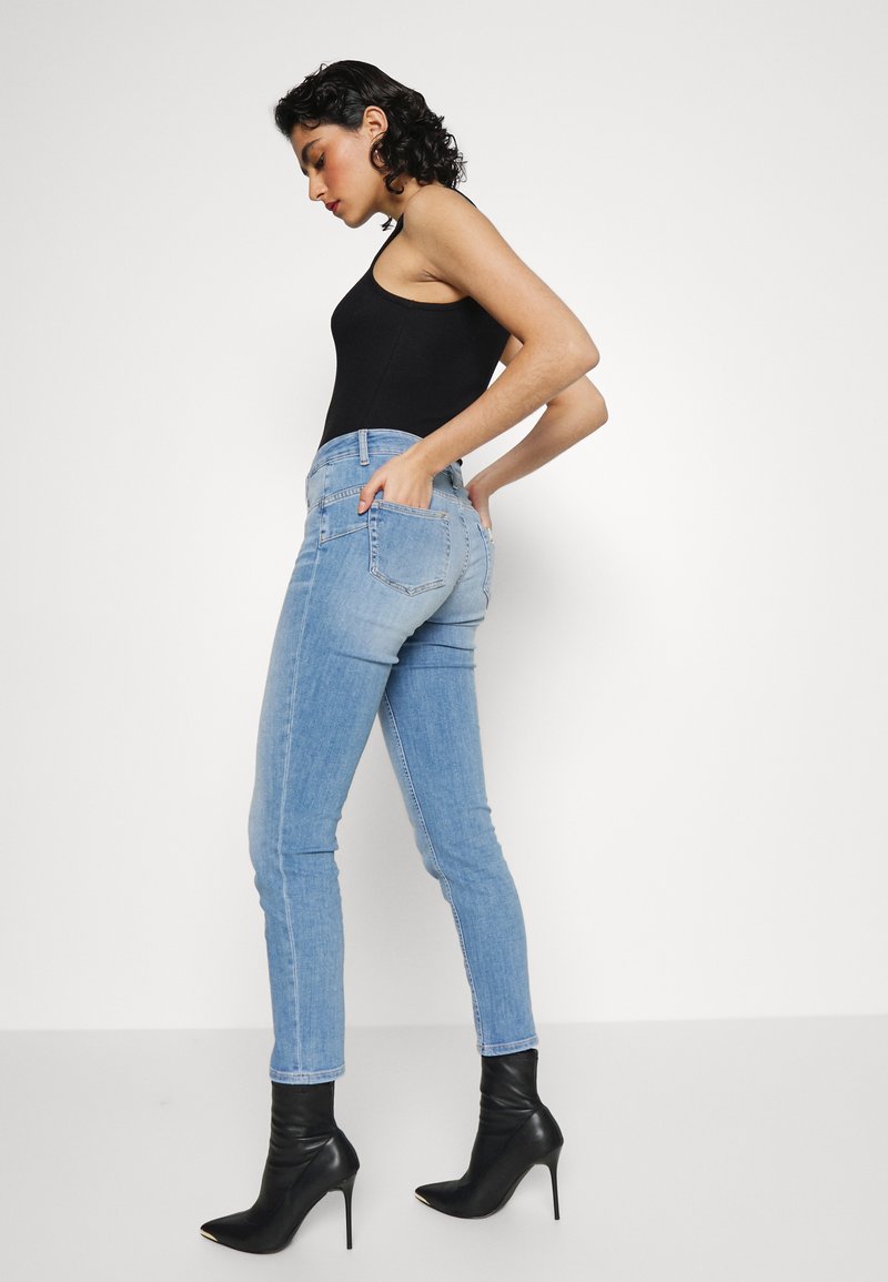 Frau in schwarzem ärmellosem Top, hellblauen engen Jeans und schwarzen hochhackigen Stiefeletten, die seitlich posiert und die Hände in den hinteren Taschen hat.