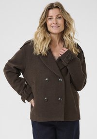 Femme aux cheveux blonds portant un manteau marron à double boutonnage avec des manches à boutons et un pantalon sombre, souriant devant un fond uni.