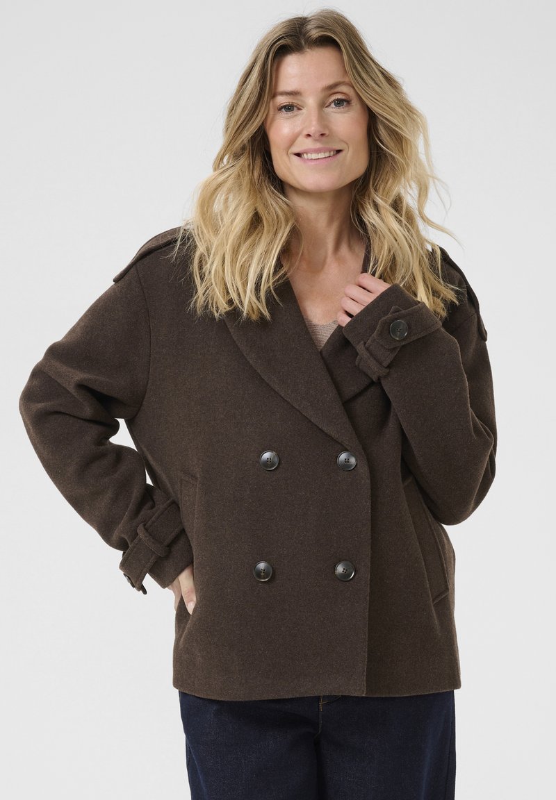 Femme aux cheveux blonds portant un manteau marron à double boutonnage avec des manches à boutons et un pantalon sombre, souriant devant un fond uni.