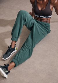 Mörkgröna fleecajoggers med elastiska muddar, kombinerade med svarta och grå sportskor med en gul accent och texturerad design.