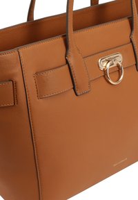 Sac à main en cuir tan avec une forme structurée, des accents en métal doré, des poignées doubles et un détail de bague décorative à l'avant.