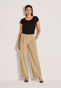 Vrouw in een zwart T-shirt met korte mouwen, beige hoge taille wijde broek, zwarte hakken met gouden details en gouden creolen, staande tegen een witte muur.