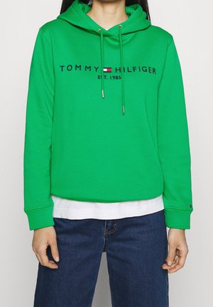 Zielona bluza z kapturem z logo "Tommy Hilfiger Est. 1985", ze sznurkami, noszona na białej koszuli i zestawiona z niebieskimi jeansami.