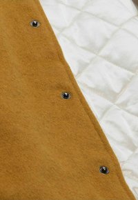 Brauner Woll-Außenstoff mit gesteppter cremefarbener Fütterung und Druckknöpfen zum Verschließen entlang des Randes. Weiche Textur mit klar gestaltetem Design.