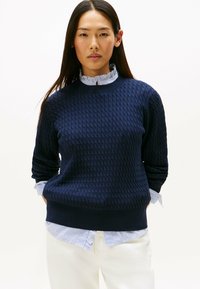 Navy blue knitted sweater with a textured pattern, round neckline, and long sleeves; layered over a white shirt with blue stripes.