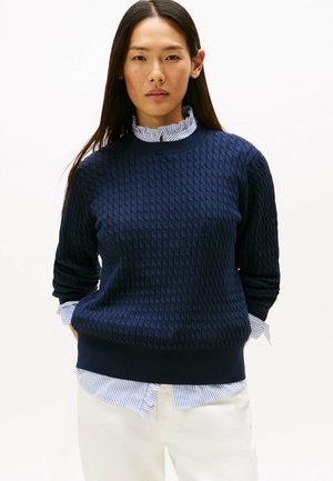 Navy blue knitted sweater with a textured pattern, round neckline, and long sleeves; layered over a white shirt with blue stripes.