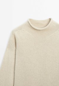 Pull beige en maille avec un col montant roulé et des manches longues, posé à plat sur un fond blanc.