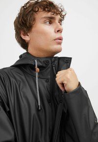 Must musta jope kõrge krae, lukuga kinnitamine ja dekoratiivsed nöörid. Läikiv tekstuur, minimalistlik disain ja nööpdetailid.