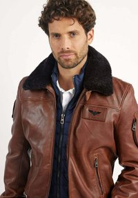 Veste en cuir marron avec un col noir en peluche, dotée de poches zippées et d'une doublure intérieure en bleu matelassé, affichant un design lourd et texturé.