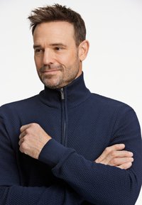 Marinblå zip-tröja med texturerad tyg, hög krage och ribbade manschetter. Modellen står med armarna i kors och visar upp det intrikata mönstret.