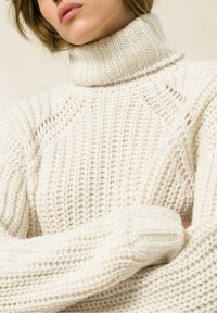 Flauschiger weißer Strickpullover mit hohem Rollkragen, gerippten Strukturen und offenen Strickdetails. Er verfügt über lange Ärmel und einen lässigen Schnitt.