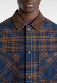 Rutiga jackan har en krage i blå och brun cordjacka, tryckknappar och två framfickor med ett rutig mönster.