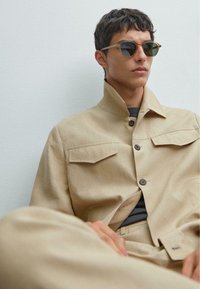 Beige Leinenjacke mit Knopfleiste und Brusttaschen, kombiniert mit einem schwarzen Hemd und runden Sonnenbrillen. Glatte Textur und taillierte Passform.