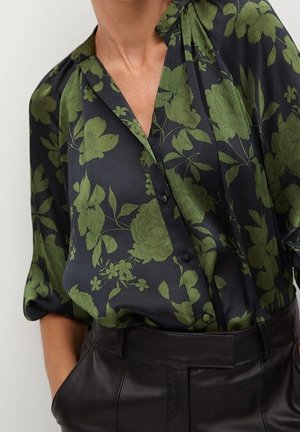 Sort skjorte med knapper og grønt blomsterprint, stoppet ned i sorte læderbukser med høj talje, med hænderne i lommerne.