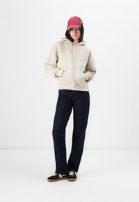 Sweatshirt beige à capuche avec poche avant, associé à un jean droit bleu foncé. Baskets noires avec accents blanc cassé et une casquette rose.