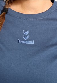 Modré tričko z měkké látky s kulatým výstřihem. Na hrudi je světle modré logo "hummel" a vzor šipek.