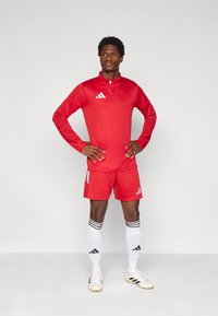 Κόκκινο αθλητικό μπλουζάκι με μακριά μανίκια και φερμουάρ στη μέση, συνδυασμένο με κόκκινο σορτς. Άσπρες ρίγες σε μαύρες κάλτσες και άσπρα παπούτσια ποδοσφαίρου με μαύρες λεπτομέρειες.