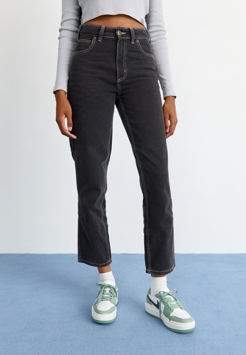 Vaqueros de mezclilla negros de talle alto con costuras blancas en contraste, con un corte recto y bolsillos delanteros, combinados con zapatillas verdes.