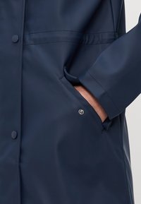 Tmavě modrý plášť do deště z texturovaného materiálu, vybavený zapínáním na patentní knoflíky, bočními kapsami a nastavitelnými manžetami. Hladký povrch a lehký design.