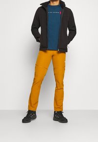 Muž v hořčicově žlutých outdoorových kalhotách, černých turistických botách, modrém tričku a černé bundě na zip, stojící před jednobarevným pozadím.