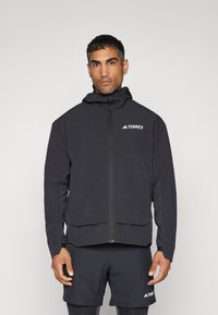 Chaqueta con capucha negra y cremallera frontal completa, hecha de un material ligero. Presenta el logo "TERREX" en el pecho; diseño minimalista.