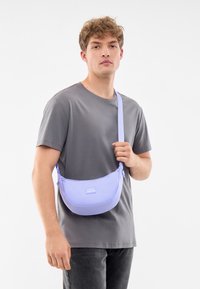 Borsa a spalla di colore viola chiaro realizzata in materiale morbido, con una forma curva e una tracolla regolabile, tenuta da una persona in t-shirt grigia.