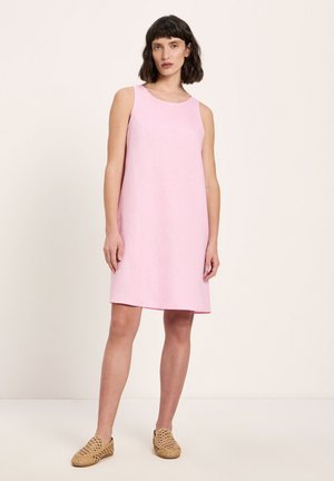 Frau steht und trägt ein ärmelloses, hellrosa knielanges Kleid sowie gewebte beige Slip-On-Schuhe vor einem einfarbigen hellen Hintergrund.