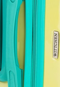 Valise rigide jaune avec fermeture éclair et poignée turquoise. Présente une plaque logo métallique marquée "WITTCHEN." Texture lisse, design moderne.