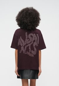 T-shirt oversized cor de vinho com um grande logótipo ondulado "ALLSAINTS" numa cor contrastante nas costas. Material macio de algodão, mangas curtas.