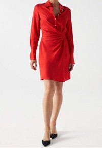 Robe rouge à manches longues avec un col, design croisé à la taille et détails de boutons. Le tissu semble lisse et léger.