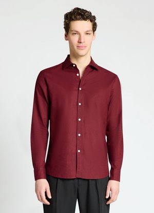 Giovane uomo con camicia bordeaux a maniche lunghe abbottonata e pantaloni neri, in piedi davanti a uno sfondo chiaro e uniforme.