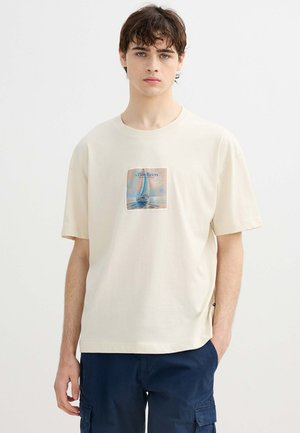 Joven con pelo oscuro que lleva una camiseta crema con un gráfico de un velero y pantalones cortos cargo azul marino, de pie sobre un fondo blanco.